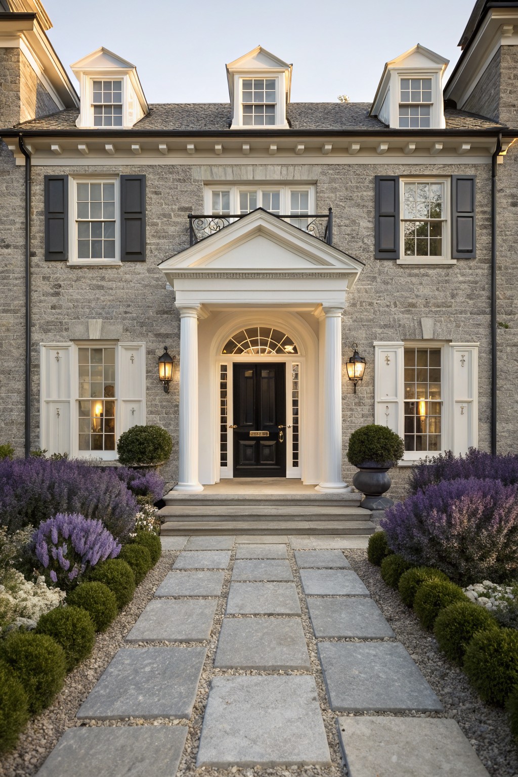 Front exterior of a two-story gray stone house with white trim, dormer windows, and a white portico supported by columns framing a black door, with lanterns, boxwood shrubs, lavender plants, and a stone pathway.