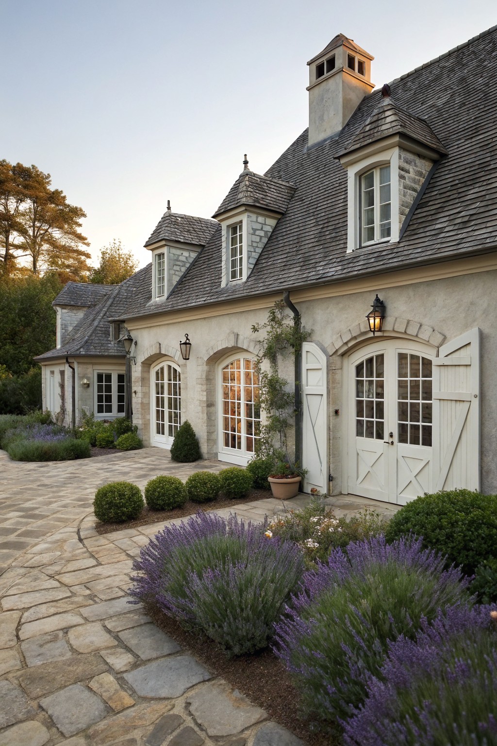 Gray stucco house exterior featuring arched white garage doors, white-trimmed windows, stone details, slate roof, lanterns, and a curved stone pathway with lavender bushes and boxwoods.