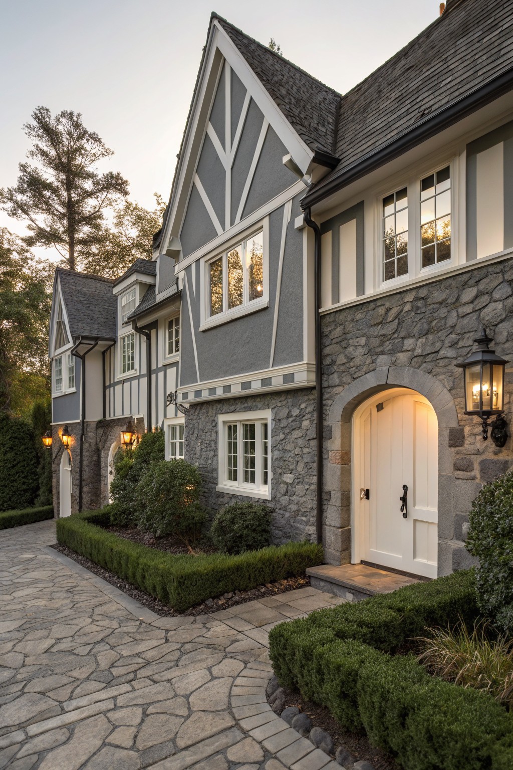 Gray shingle and clapboard house exterior with white timber framing and trim, stone base wall, arched white entry door with lanterns, boxwood hedges, and irregular stone paver driveway in evening light.