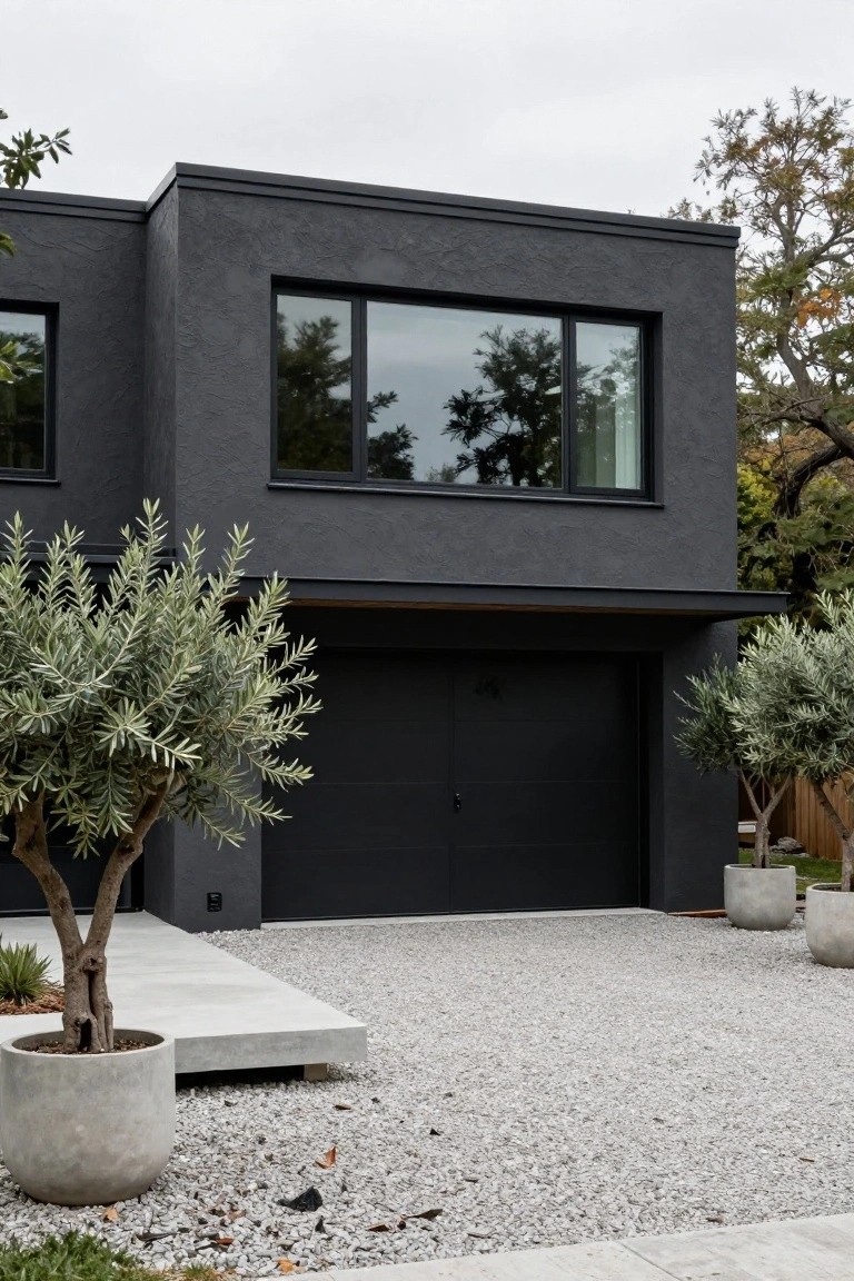 Modern house exterior with textured charcoal gray walls, large black-framed windows, black garage door, potted olive trees, and gravel driveway.