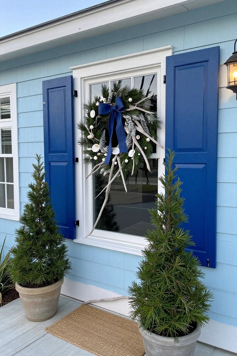Light blue clapboard house exterior with dark blue shutters framing a window hung with a Christmas wreath of pine boughs, white shells, driftwood branches, pinecones, and a large navy bow, flanked by two small potted evergreen trees in terracotta and gray pots, a lit black lantern on the wall, and a seagrass mat on the porch.