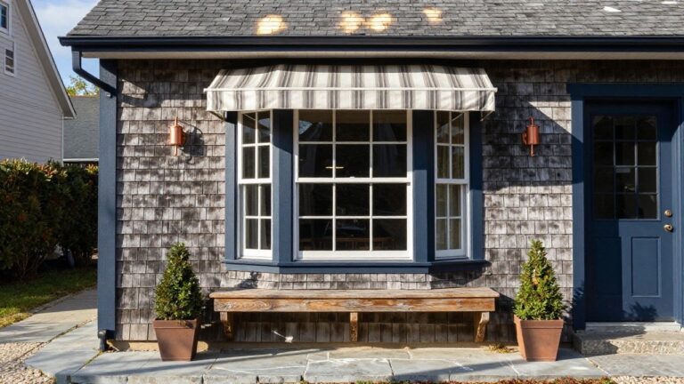 Front view of a shingle-clad house exterior with a central bay window framed in navy blue, flanked by copper lanterns, a wooden bench below the window, boxwood shrubs, and a stone pathway leading to steps.