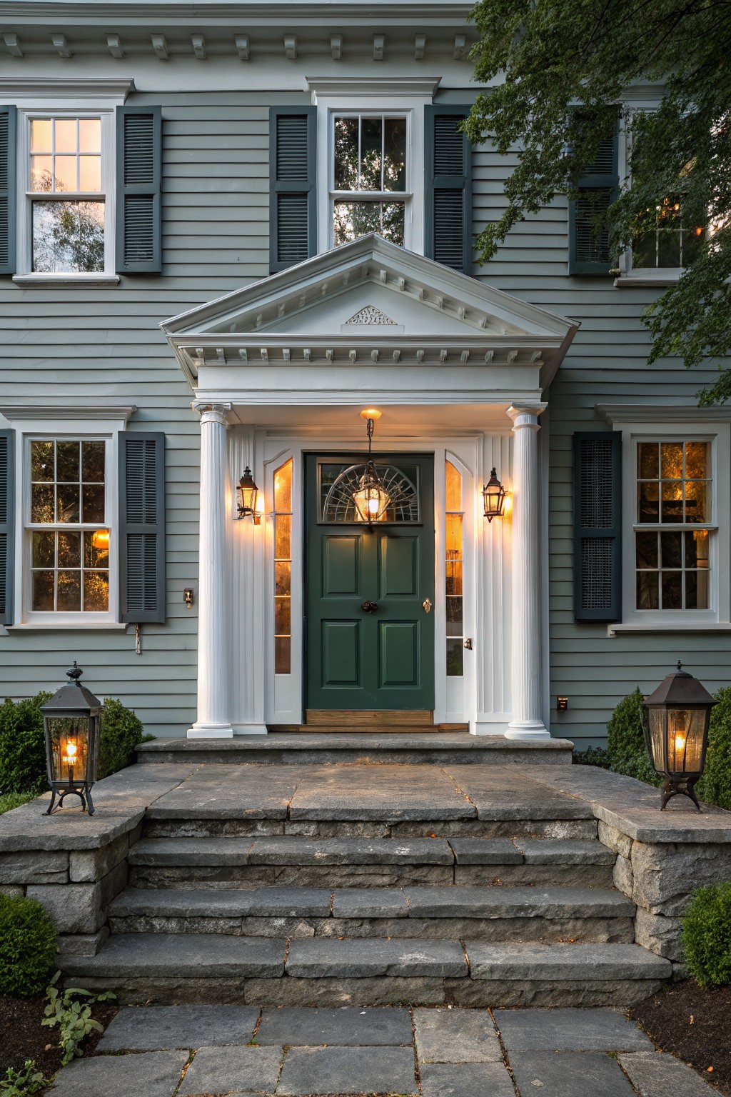 Gray clapboard house facade with white columns framing a green front door, lanterns on porch and steps, flanked by shrubs and stone stairs.
