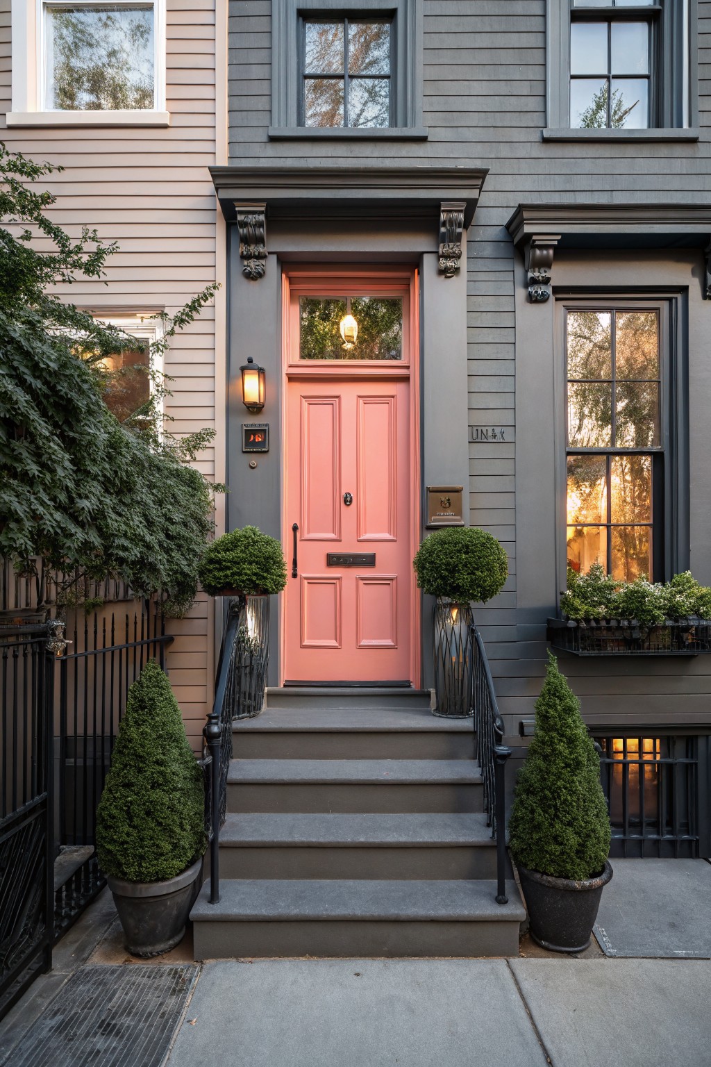 Gray rowhouse facade with pink double front door, flanked by potted topiary shrubs on stone steps with black iron railing, lantern light, and window box planting.