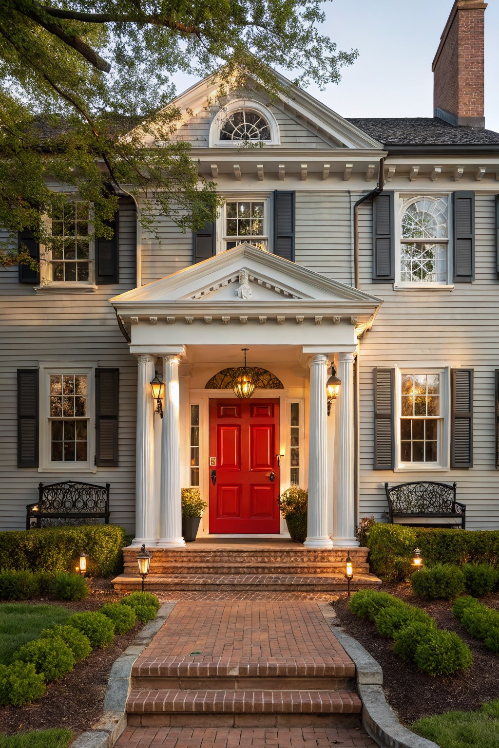 Gray shingle-sided house exterior with white-columned portico, red double front door, black shutters, lanterns, brick steps and path, boxwoods, and evening lighting.