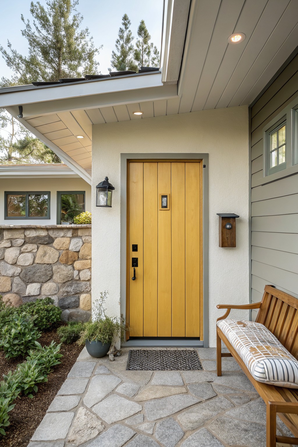 Yellow wooden front door with black hardware on a light stucco wall next to gray siding, with a stone retaining wall, wooden bench, potted plants, and flagstone path leading up to it.