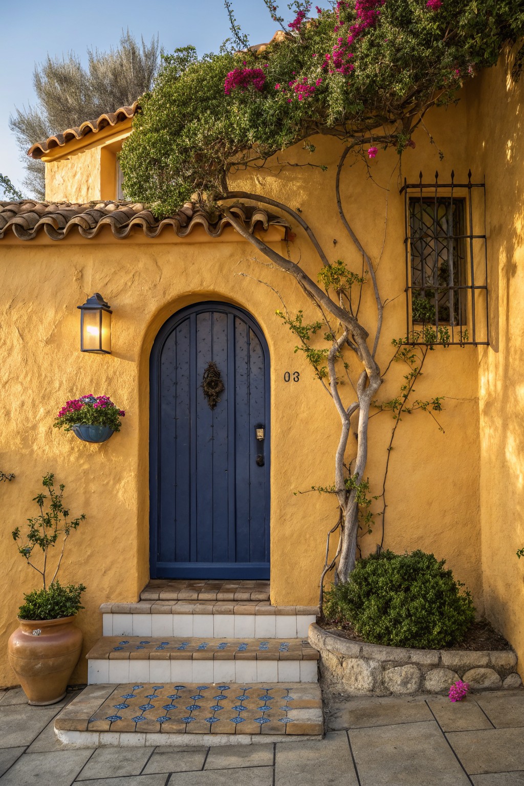 Yellow stucco house exterior with terracotta tile roof, arched navy blue wooden door numbered 03, wrought iron window grille, hanging lantern, blue flower basket, climbing vines and bougainvillea, potted plants, tiled steps, and stone base.