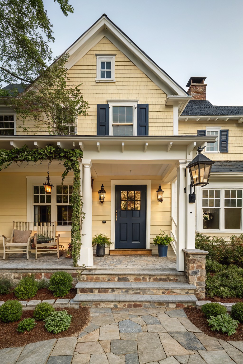 Pale yellow shingle-style house with white trim, navy blue front door, covered porch supported by columns, hanging lanterns, Adirondack chairs, potted plants, and stone steps leading to a bluestone pathway edged with shrubs.