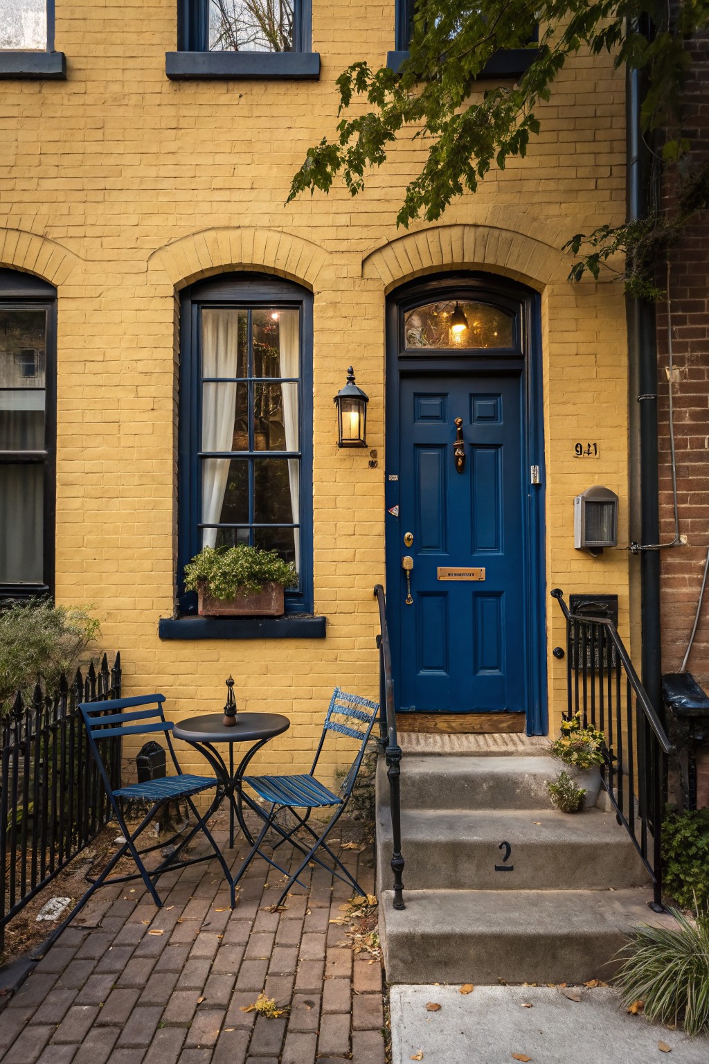 Yellow brick rowhouse exterior featuring a navy blue front door with brass knocker, arched transom window, black lantern light, window box planter, small front patio with bistro table and chairs, black iron fence, and adjacent brick building.