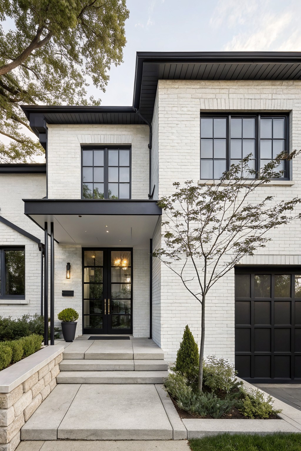 Two-story white brick house exterior with black metal trim, black-framed windows, glass front door under a cantilevered black canopy, black garage door, concrete steps to the entry, and minimal landscaping including a small tree and shrubs.