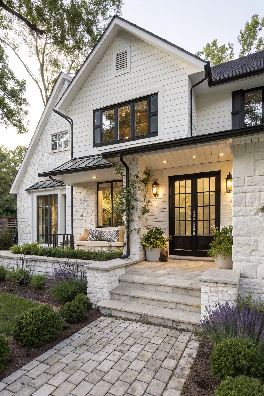 White clapboard house exterior with black-framed windows and double doors at the entry, metal awning over porch, stone pillars, steps leading to brick paver path, and low plantings along the front.