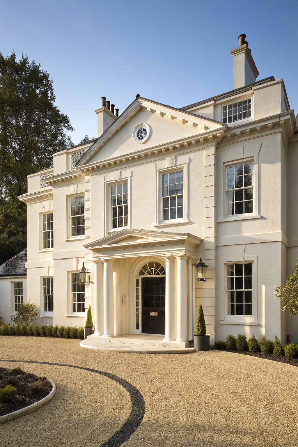Two-story white stucco house with symmetrical Georgian facade, columned portico entrance with lanterns, sash windows, gravel driveway, and evergreen shrubs.