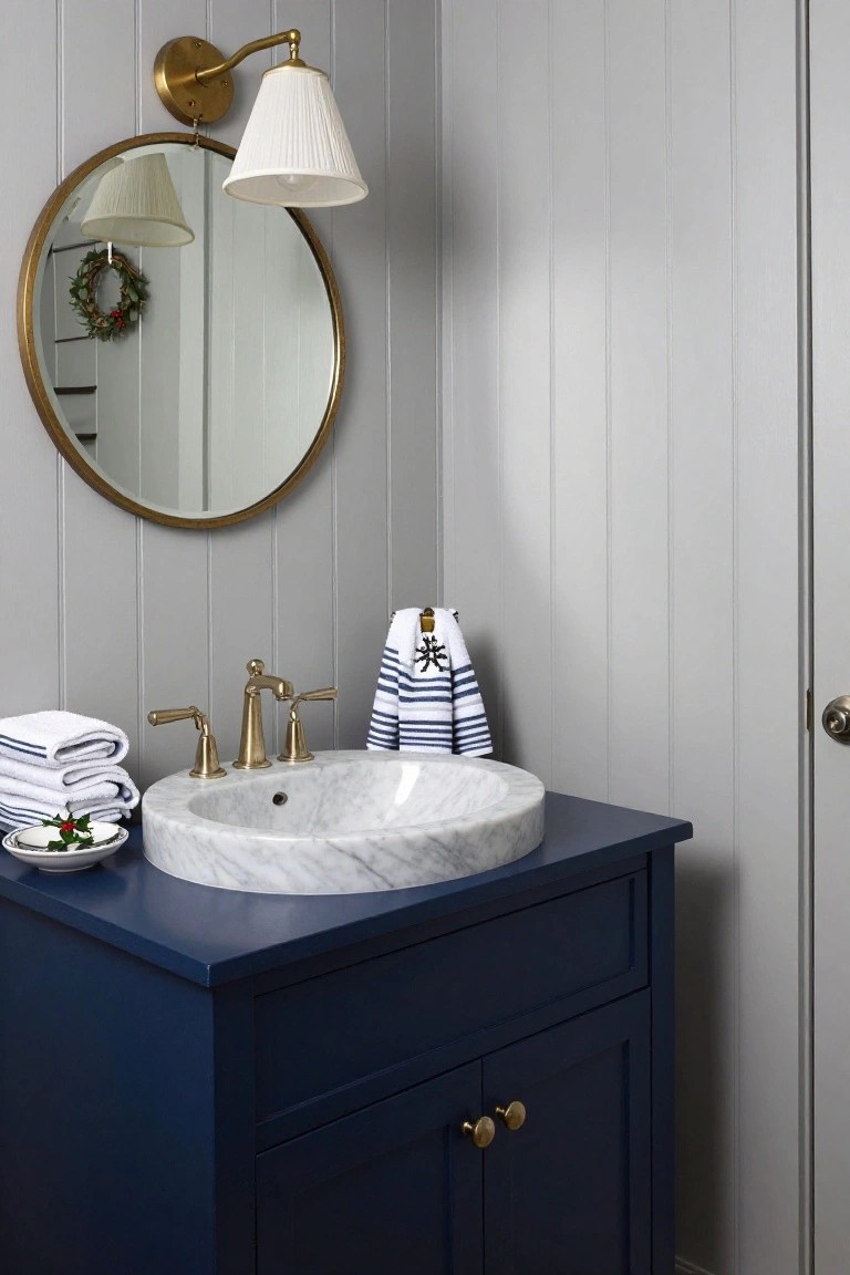 Navy blue wooden vanity with white marble vessel sink, brass faucet and pulls, stacked white towels and navy striped towels with nautical motifs, small plates with holly sprigs, round brass mirror with wreath, white lamp on gray shiplap wall in a bathroom.