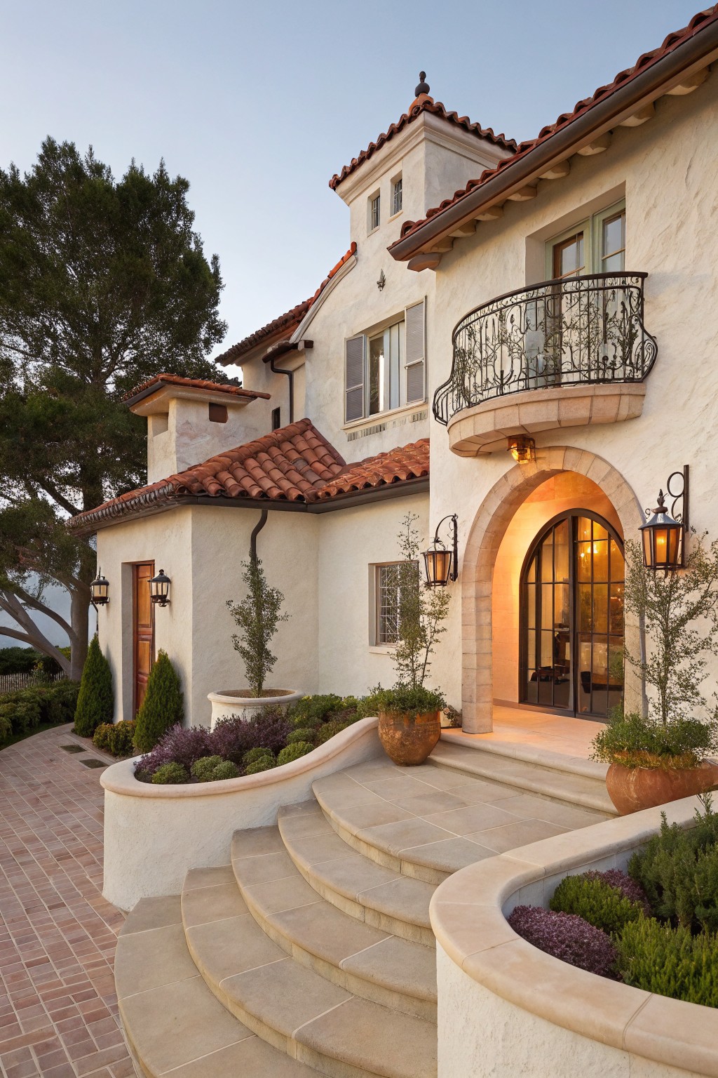 White stucco house exterior with red tile roof, curved beige stone steps leading to an arched entry with black-framed glass doors, wrought iron balcony above, wall lanterns, potted plants, and surrounding trees and shrubs.