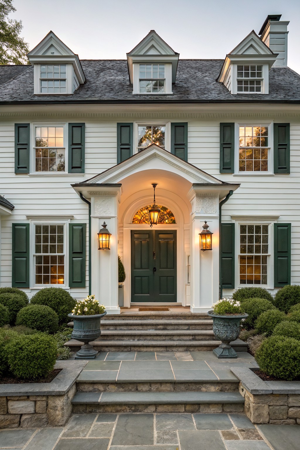 White two-story house with gabled roof, dark green shutters, and a central white portico sheltering a dark green door, accessed by wide bluestone steps with stone retaining walls and potted shrubs on either side.