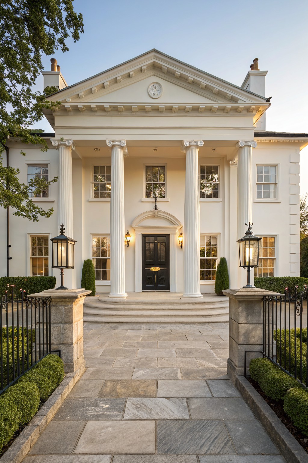 White neoclassical house facade with four tall fluted columns, pediment, clock, black front door, stone piers with black wrought iron gates and railings, stone steps, lanterns, and boxwood shrubs.