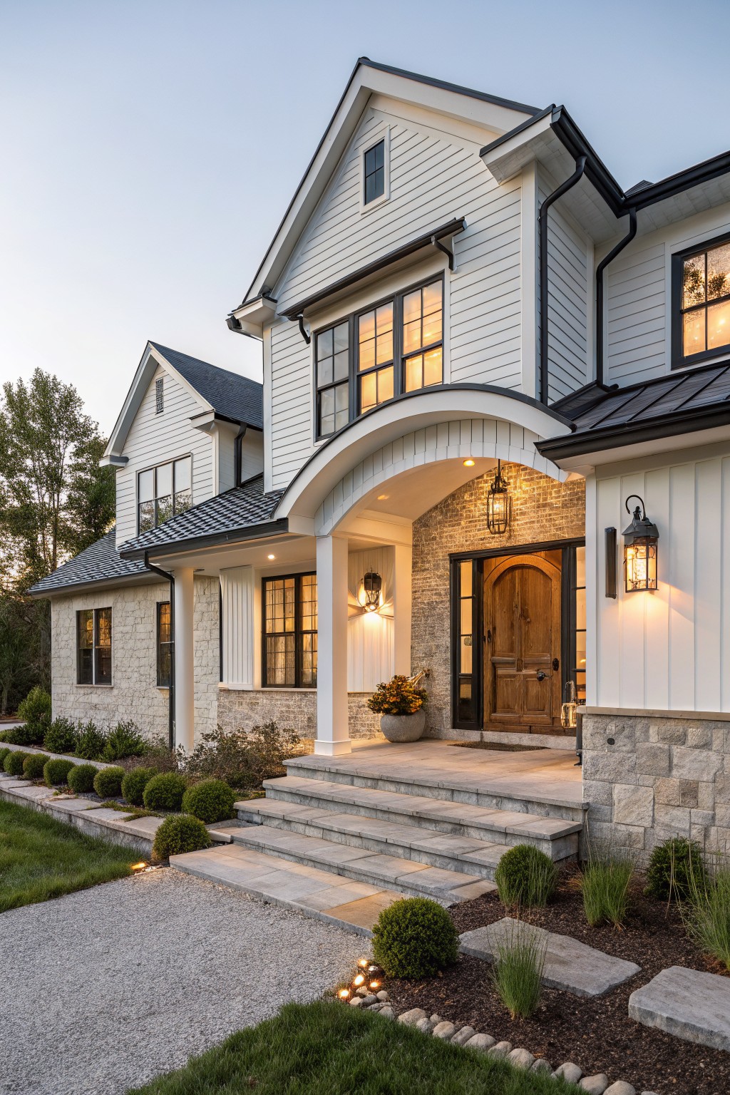 White shiplap house exterior with black metal roof, arched covered porch entry supported by stone pillars and base, wooden arched front door with black hardware, lanterns on posts, stone steps, boxwood shrubs, gravel driveway, and lawn at dusk.