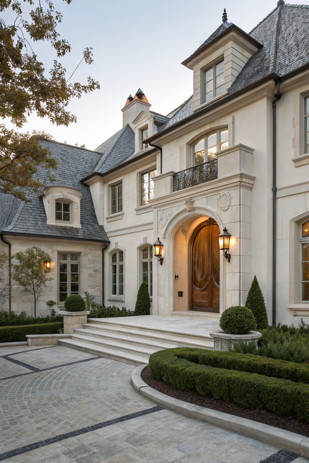 White stucco house exterior with dark slate roof, grand arched limestone entryway featuring double wooden doors and lanterns, marble steps to a circular gravel driveway edged by boxwood hedges.