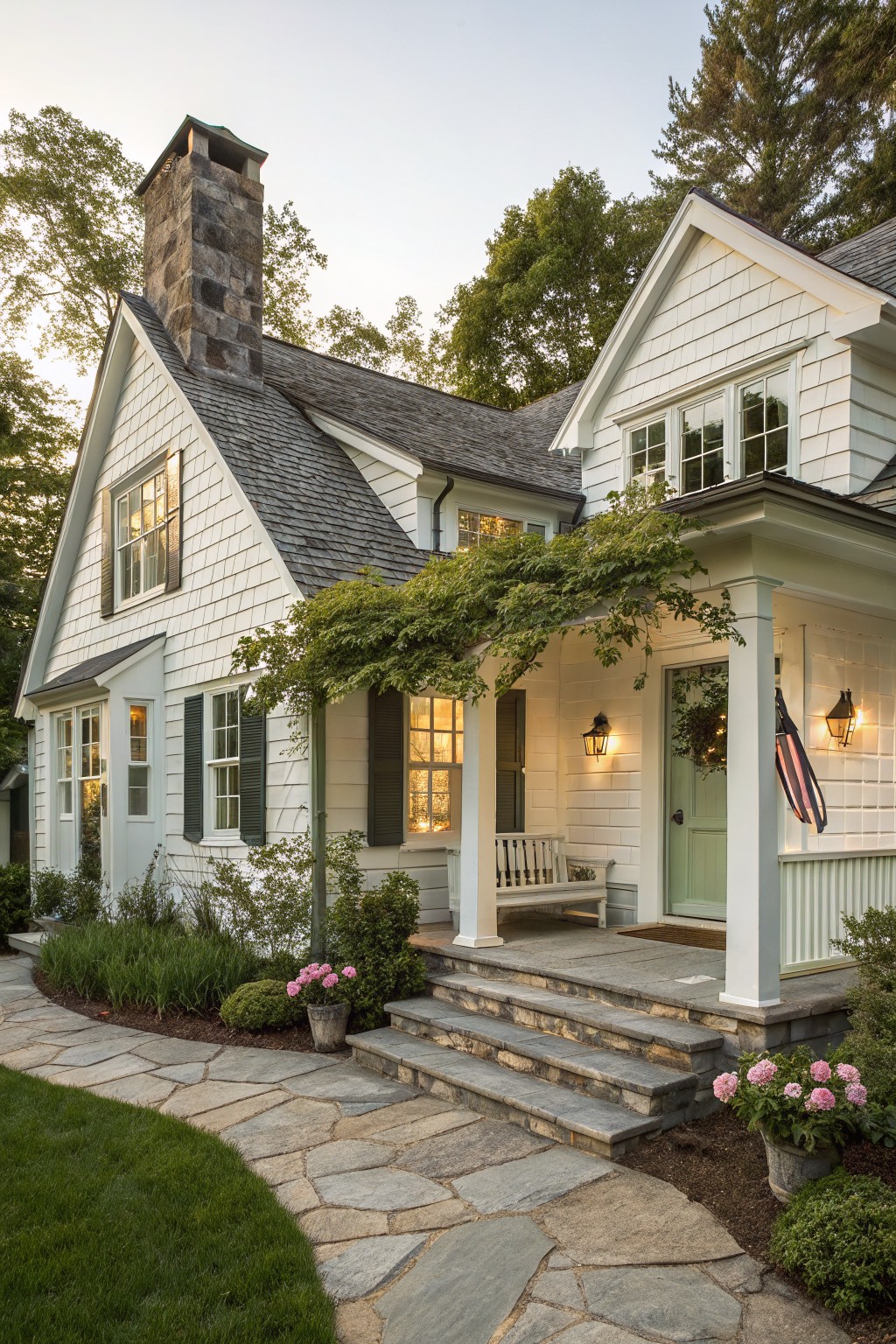 White shingle-style two-story house with tall stone chimney, covered front porch with green door and lanterns, stone steps and flagstone path, potted flowers, and surrounding greenery and trees in evening light.