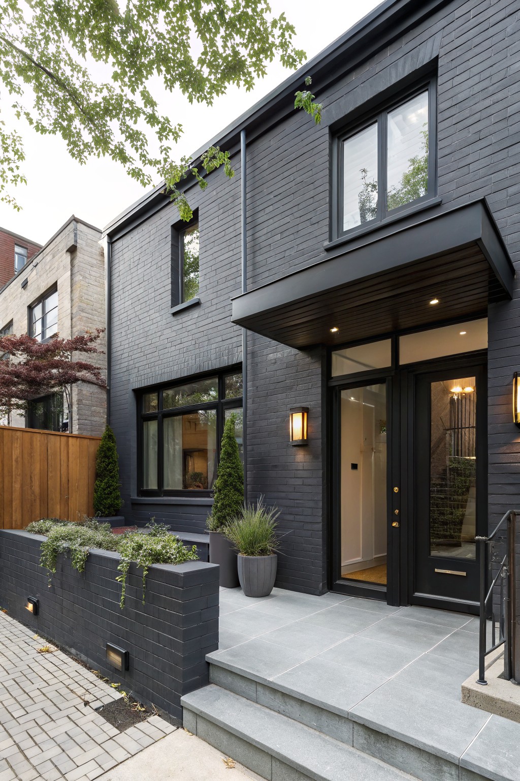 Dark gray brick two-story house exterior with cantilevered black entry canopy, glass front door, wall-mounted lanterns, brick planters with greenery, and gray stone steps.
