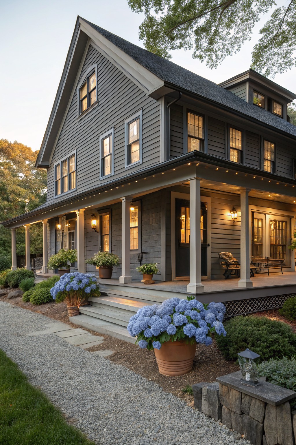 Two-story gray shingle house with wrap-around porch, white columns, lit soffit lights and lanterns, large blue hydrangea pots, stone steps, and gravel path at dusk.