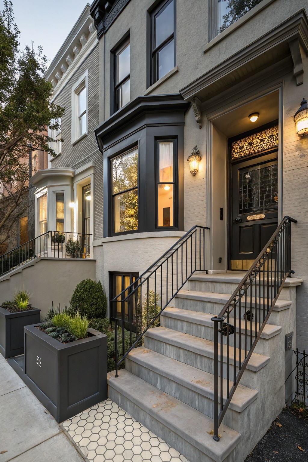 Gray brick rowhouse exterior with black-framed windows, a black front door featuring stained glass transom, wrought iron railings along concrete stoop steps, potted plants in black containers, and hexagonal tile pathway at the base.