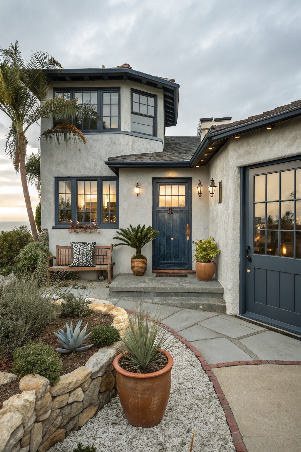 Light gray stucco house exterior featuring a navy blue front door with sidelights, matching garage door, dark-framed windows, wooden bench, potted plants, and stone pathway.