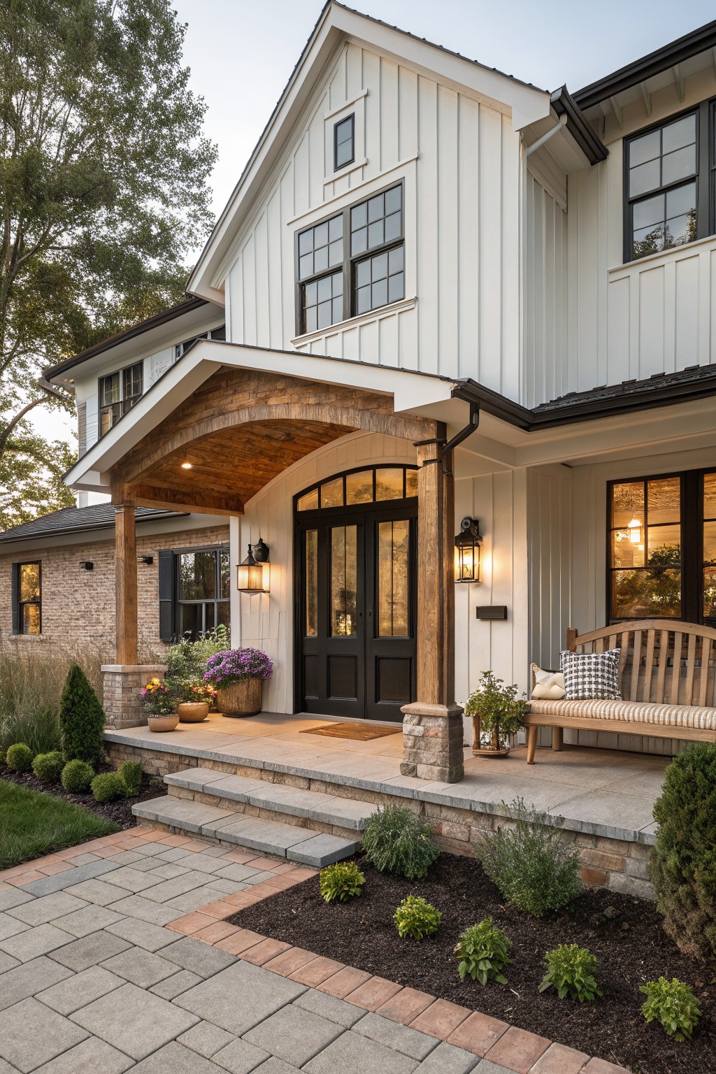 White board-and-batten sided house exterior with covered front porch supported by stone pillars, dark wood beams, black double doors, lanterns, potted plants, bench, and brick accents on the side foundation.