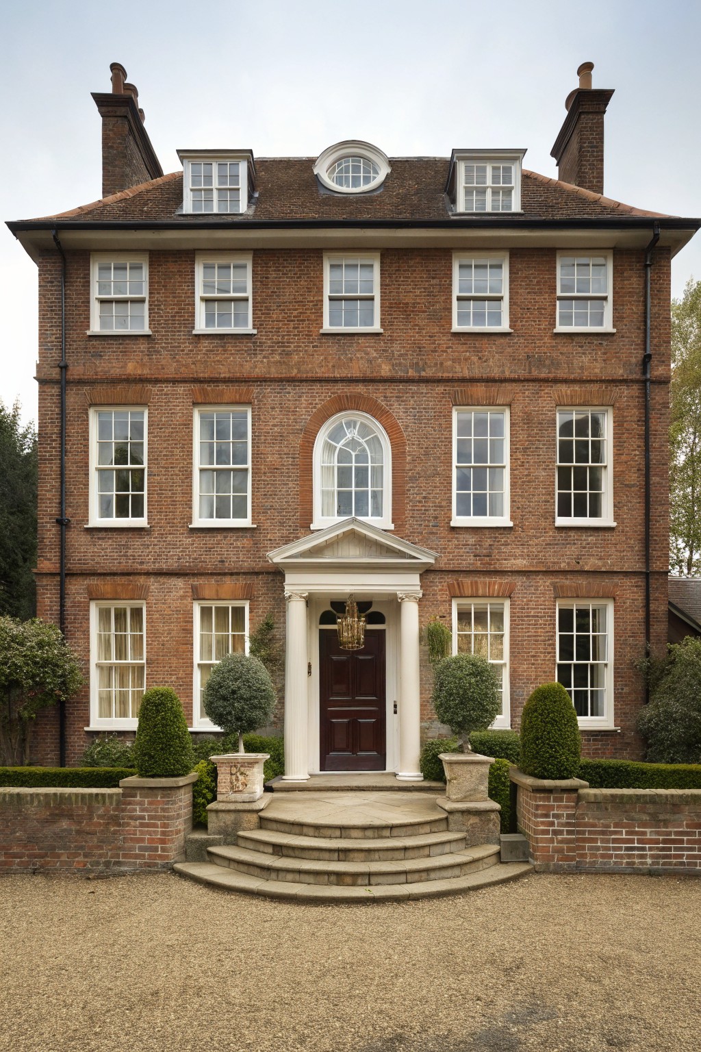 Three-story brown brick house with symmetrical white sash windows, central white pedimented portico supported by columns over a dark wood front door, flanked by lanterns and topiary shrubs on low brick walls with gravel driveway in front.