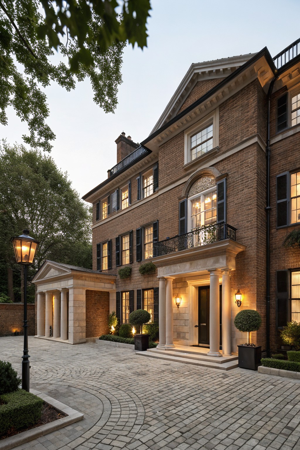 Two-story brown brick house exterior at dusk with classical stone portico entry supported by columns, flanked by lanterns, circular gravel driveway, and low landscaping.