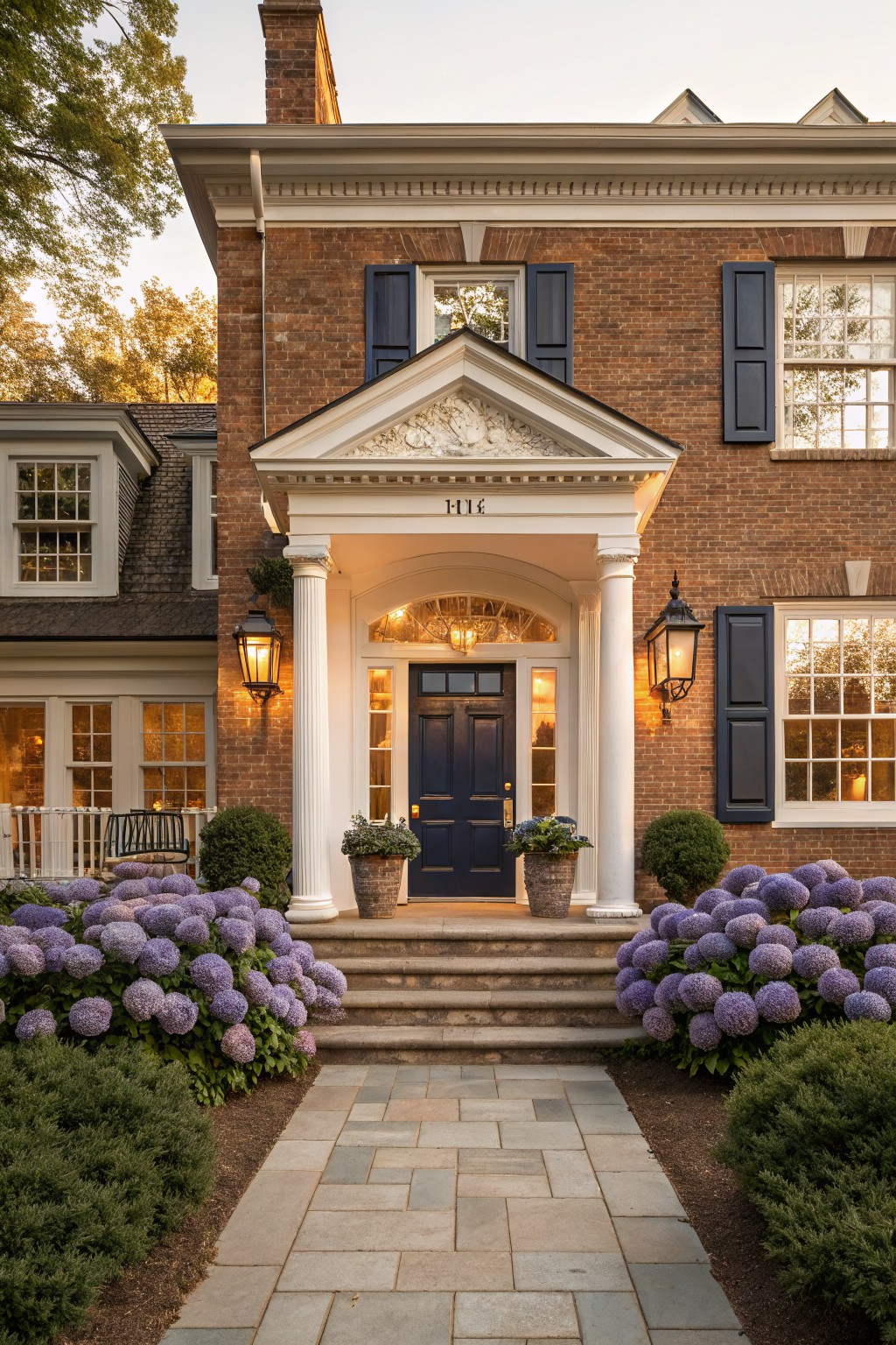 Brown brick house exterior featuring a white classical portico with columns and pediment over a navy blue front door, flanked by lanterns, steps leading to a stone pathway, and purple hydrangea bushes.