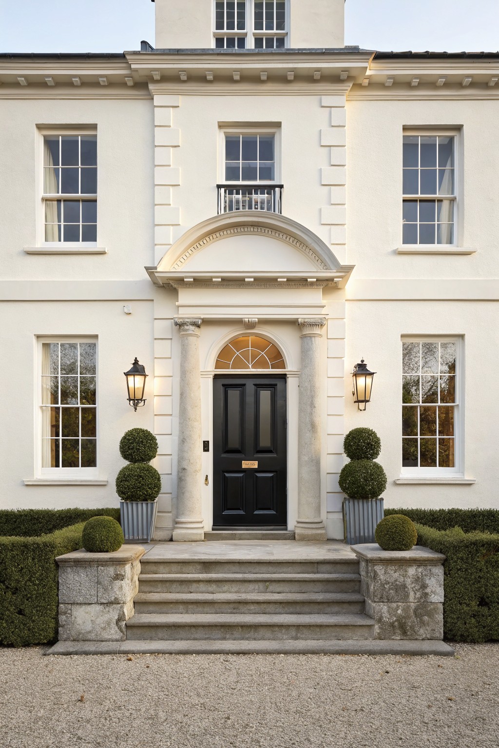 White stucco house exterior featuring a central columned portico with black front door, fanlight window, stone steps, lanterns, and paired boxwood topiaries on pedestals.