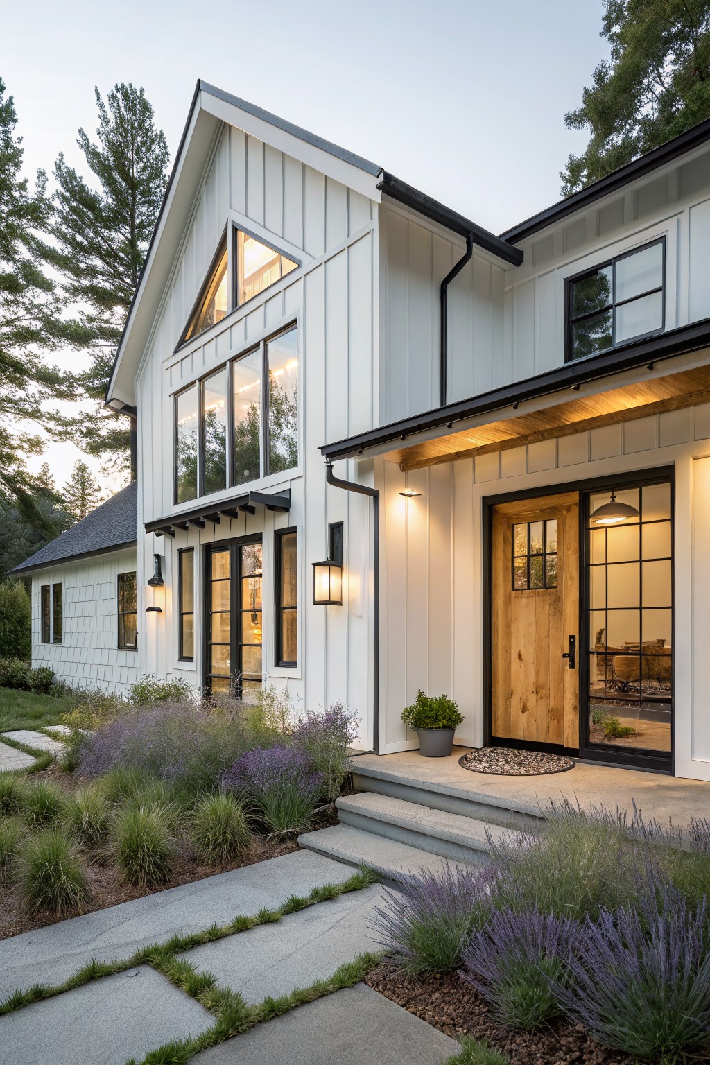White two-story house exterior with board-and-batten siding, black window frames, wooden front door with glass panels and black metal surround, porch lanterns, concrete steps, lavender plants, and stone path at dusk.