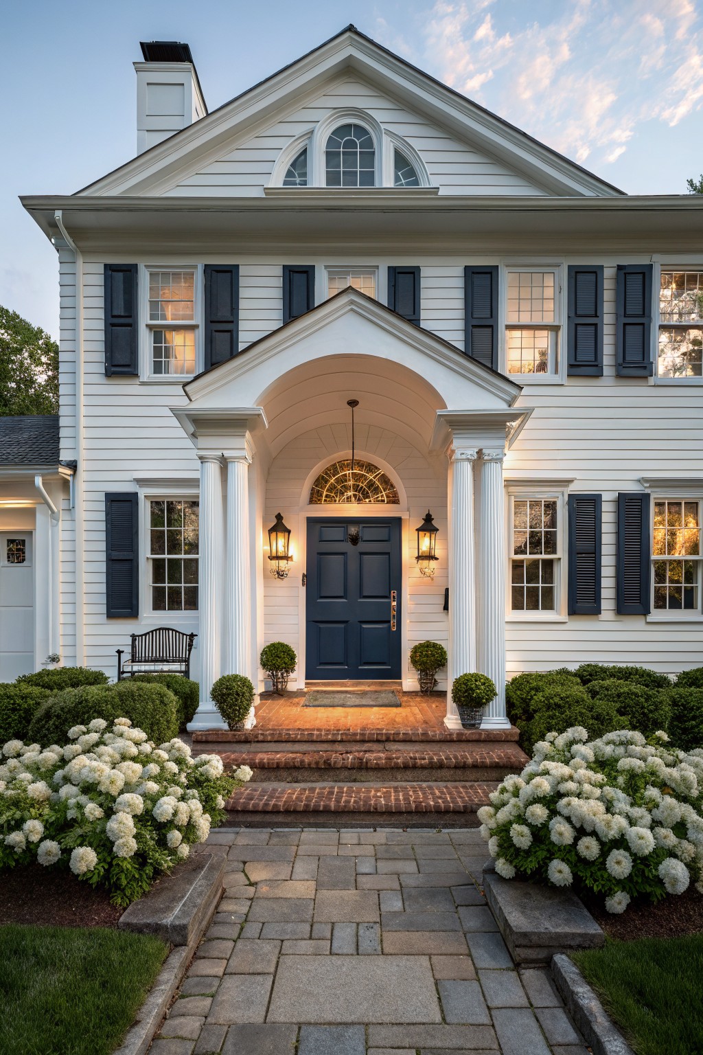 White two-story colonial house exterior with navy blue front door, matching shutters, columned portico, brick entry steps, hydrangea shrubs, and bluestone pathway at dusk.