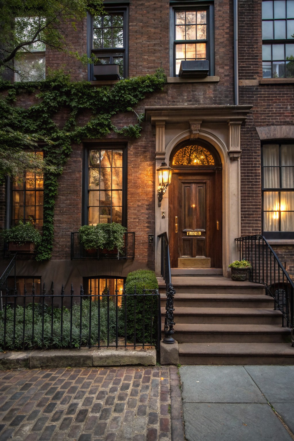 Brown brick townhouse exterior with ivy-covered walls, arched wooden front door illuminated by a wall lantern, potted plants on steps, window boxes, and warm light from windows at dusk.