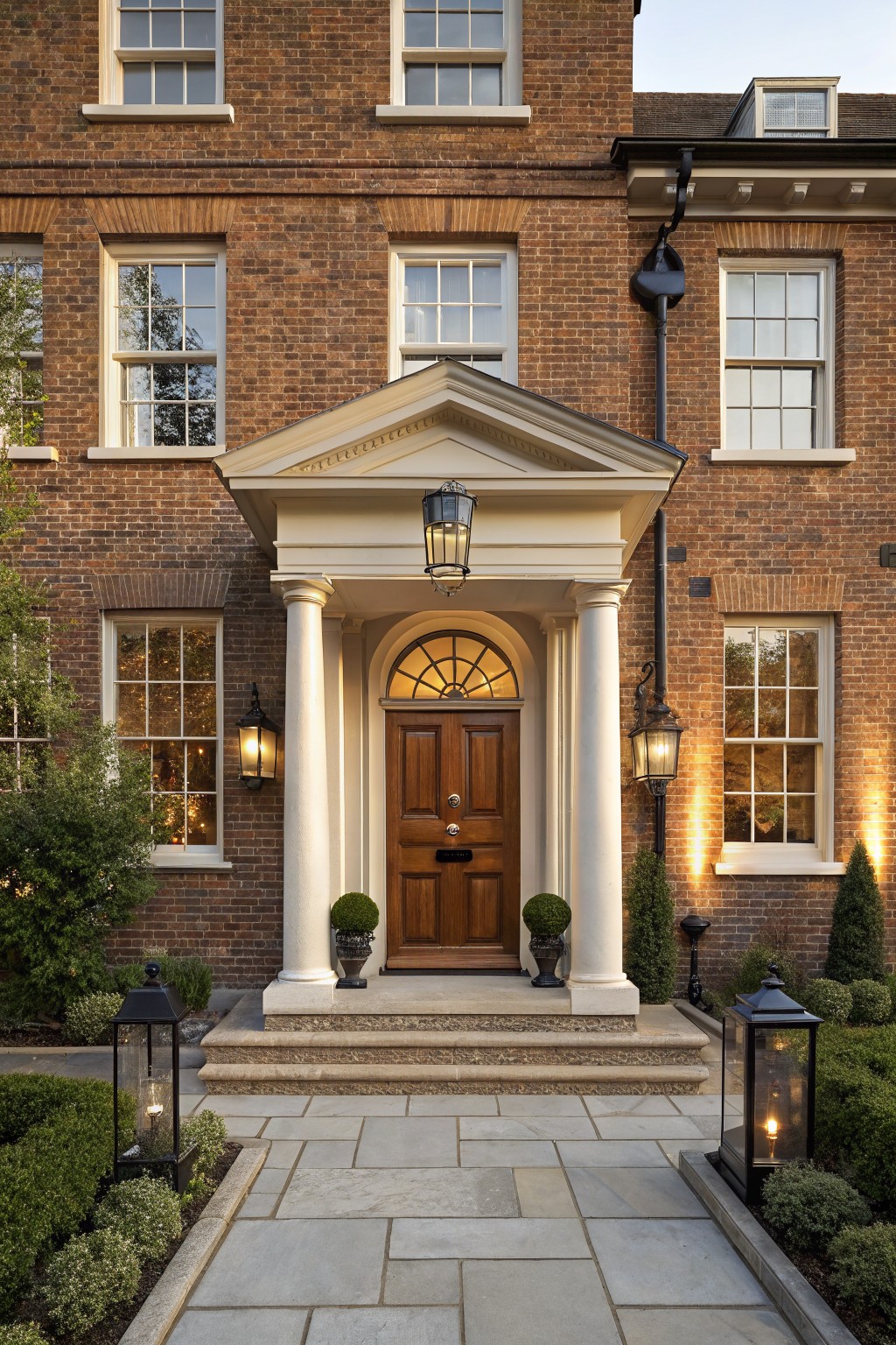 Red brick house exterior with a white neoclassical portico featuring fluted columns, a semicircular fanlight above a paneled wooden front door, wall-mounted lanterns, potted topiaries, and a stone pathway edged by boxwood shrubs.