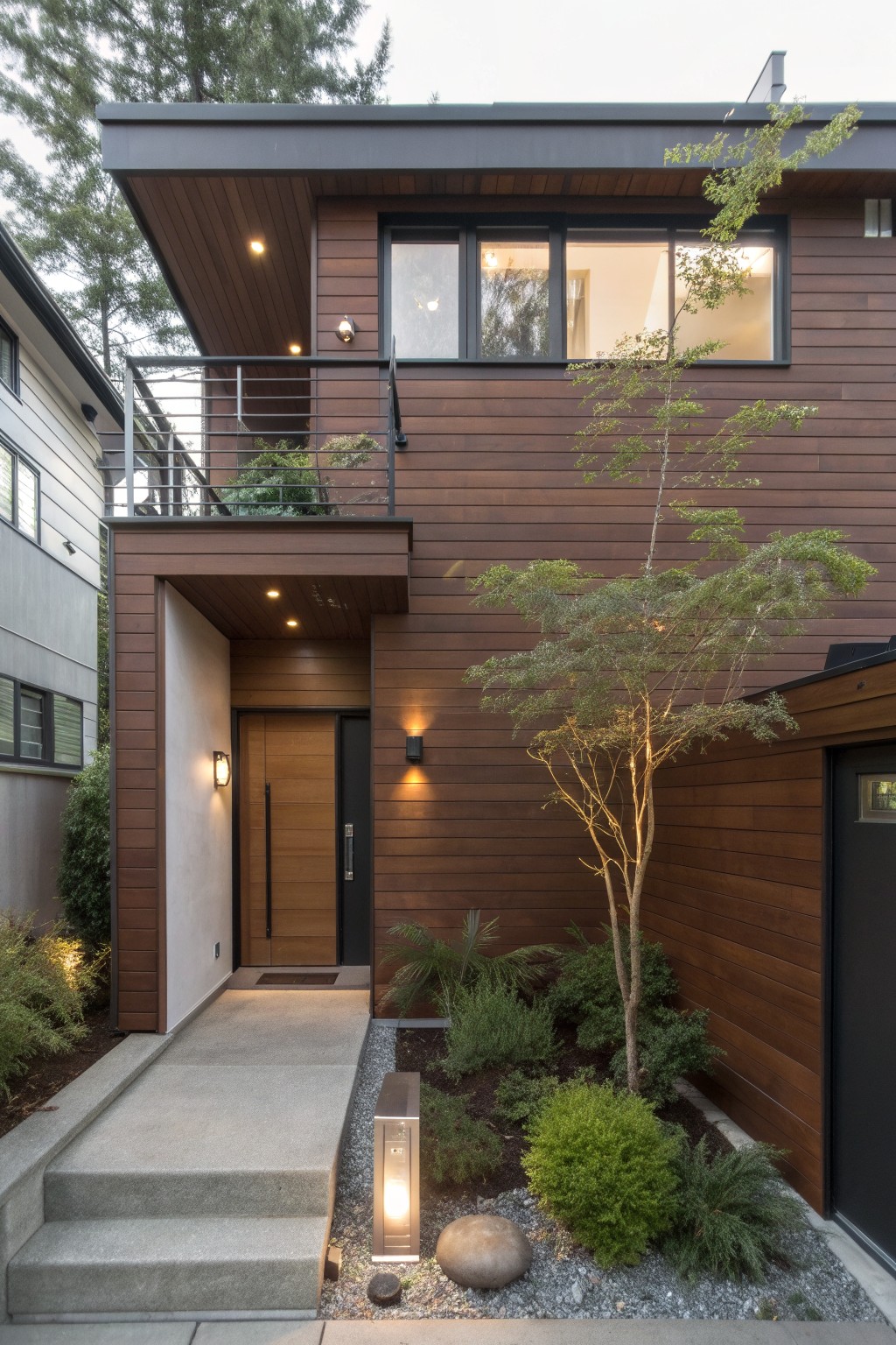 Modern two-story house exterior with warm brown horizontal wood cladding, black metal balcony railing, wooden front door, stucco entry area, and small landscaped front yard with a tree, gravel, and plants.