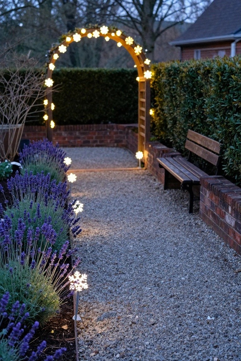 Gravel garden path lined with lavender plants and illuminated by snowflake-shaped stake lights, leading under a wooden archway wrapped in snowflake string lights, with a wooden bench against a brick wall beside hedges.