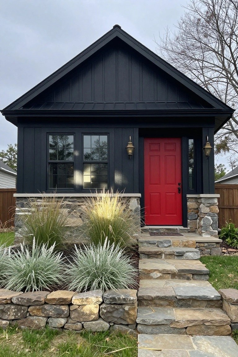 Small black board-and-batten Craftsman house exterior with red front door, black-framed windows, stone pillars, ornamental grasses, and flagstone steps on a grassy yard.