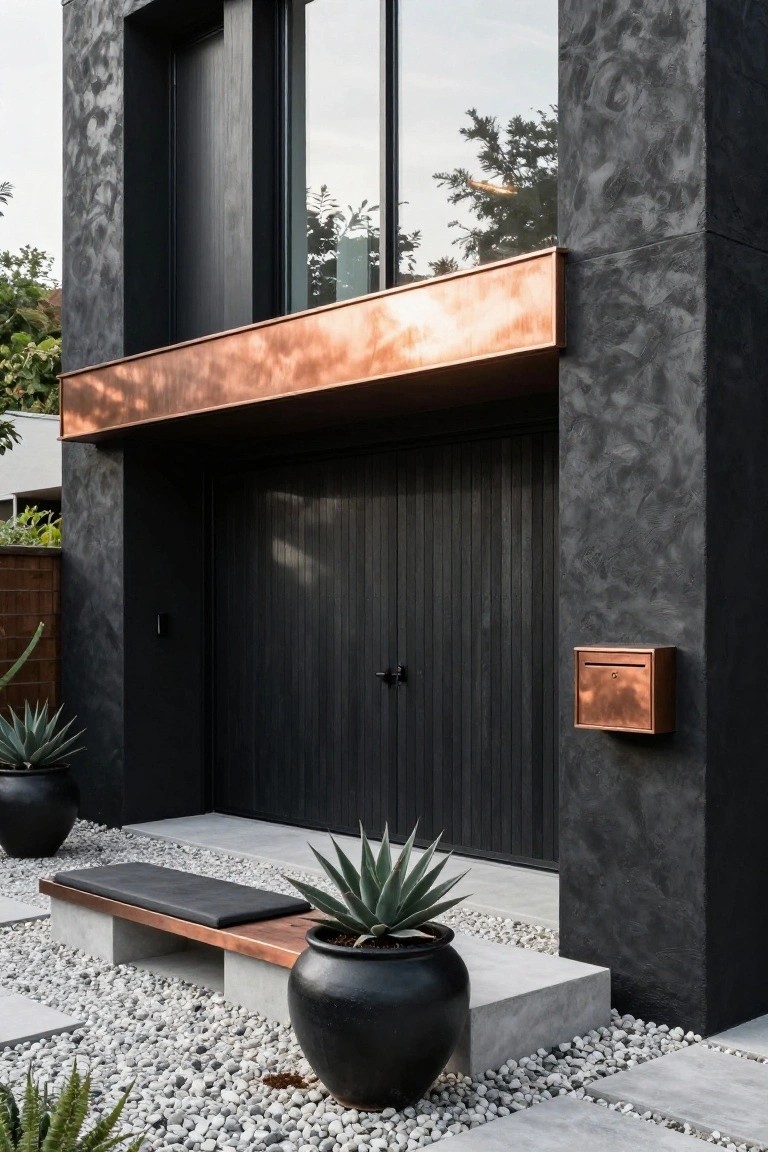 Black textured house facade with recessed copper awning over dark vertical wood garage door, copper mailbox, concrete bench, black pots with agave plants, and white gravel ground cover.