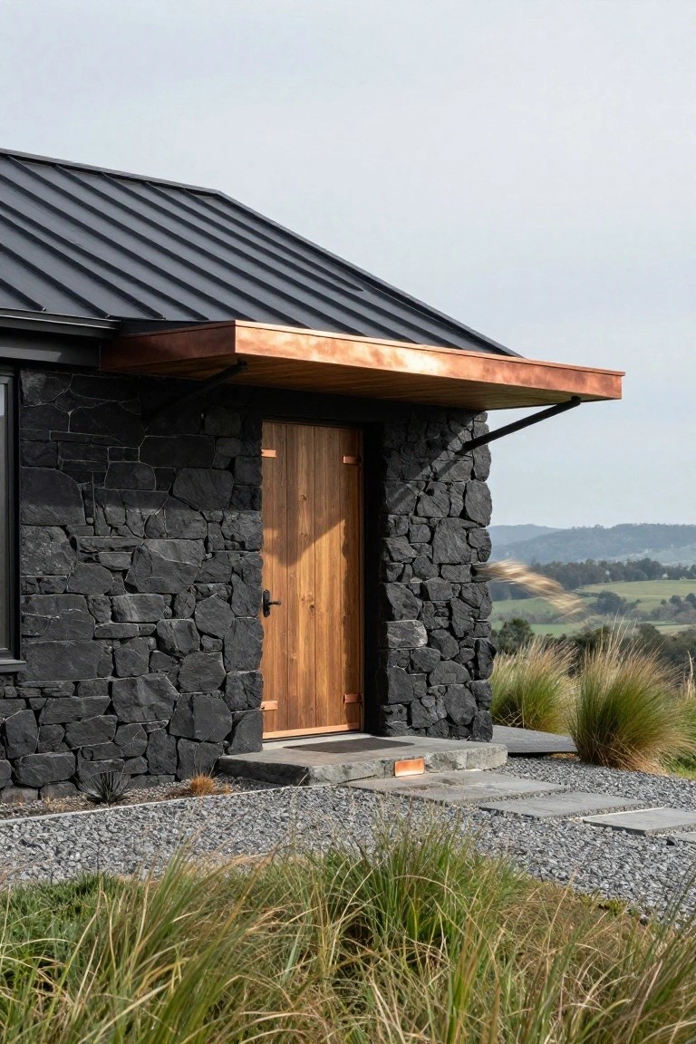 Black stone house exterior featuring a wooden entry door under a cantilevered copper awning, with gravel path, stone steps, native grasses, and rolling hills in the background.