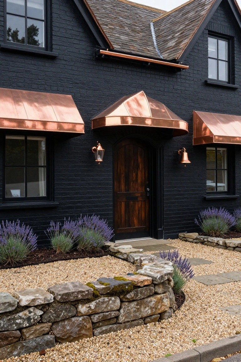 Front view of a two-story black brick house with gabled slate roof, copper awnings over black-framed windows and arched wooden door, copper lantern lights flanking the entry, purple lavender plants, gravel path, and dry-stacked stone wall bed.
