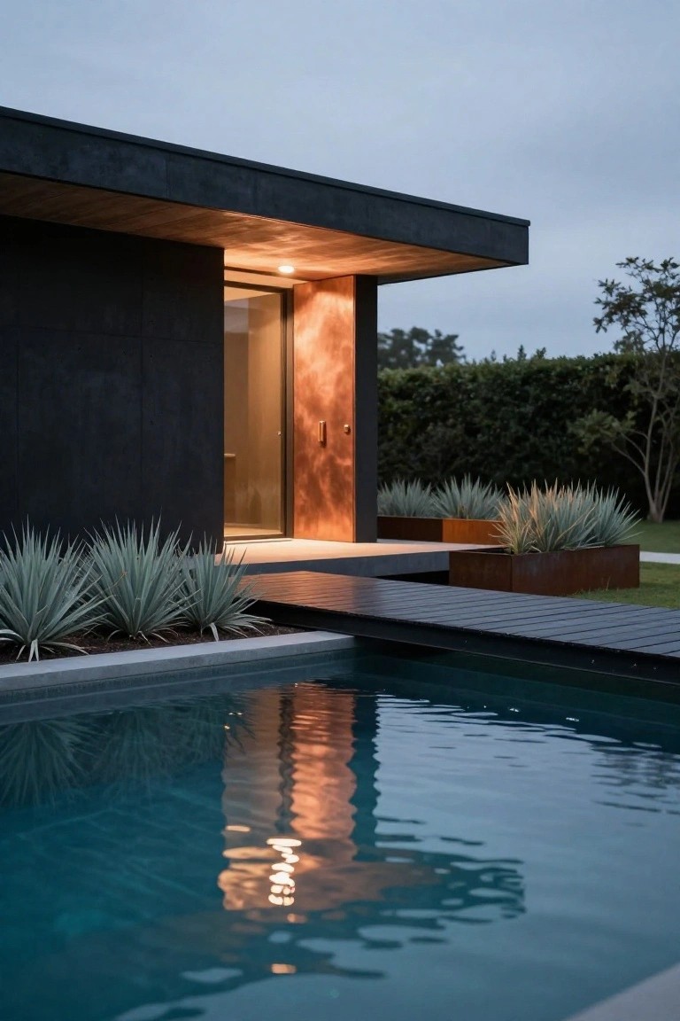 Black modern house corner with copper entry door, illuminated wooden walkway leading to turquoise pool, agave plants in corten steel planters, and hedge backdrop at dusk.