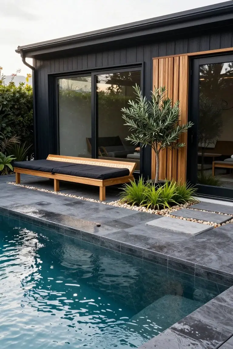 Black timber-clad modular house exterior with vertical wooden slats beside black-framed sliding glass doors, next to a pool edge, wooden daybed, olive tree, grasses, pebbles, and slate paving.