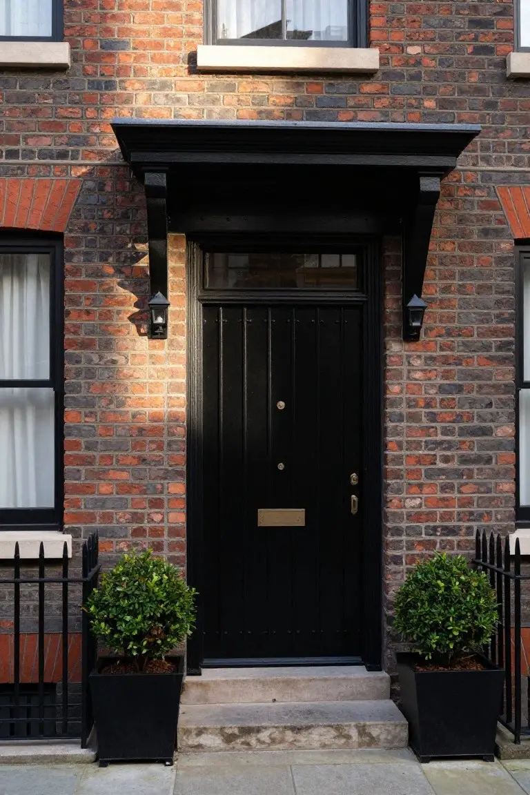 Red brick terraced house exterior with black-framed sash windows, black wood front door with brass knocker and letterbox, black lanterns flanking the door, and matching black planters with topiary on stone steps.