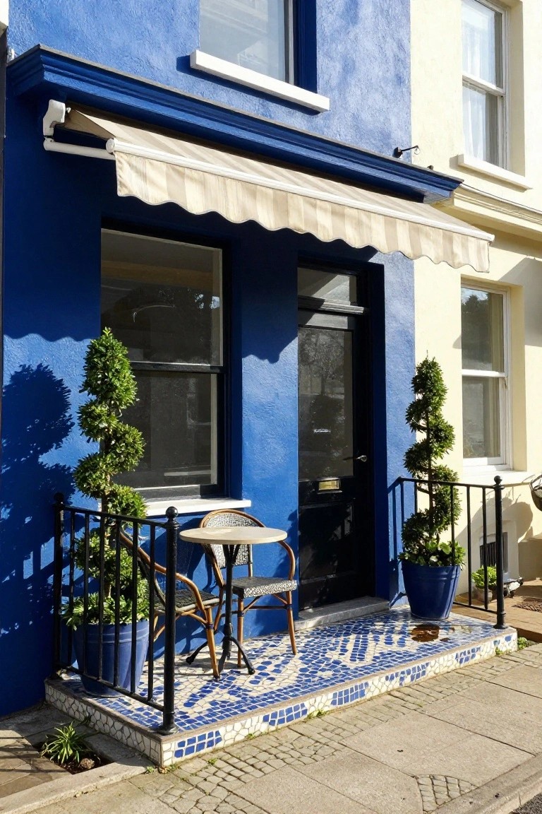 Blue painted terraced house facade with black front door under beige awning, white bistro table and two chairs on mosaic tiled porch step, iron railing with potted spiral topiary trees, adjacent pale yellow house wall.