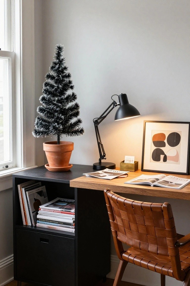 A small black artificial Christmas tree in a terracotta pot next to a wooden desk with open books, a black desk lamp, gold box, and framed abstract art in a home office with black shelving unit and brown woven leather chair.