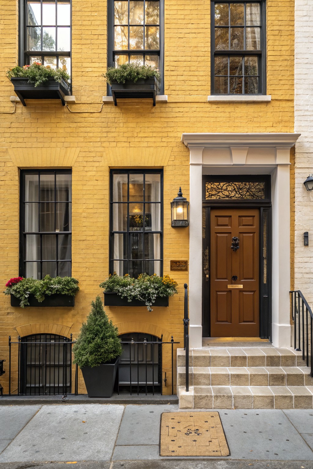 Narrow yellow brick townhouse exterior with black-framed windows, flower boxes containing plants, potted shrubs, brown wooden front door, stone steps, wrought iron railing, and tan doormat on city sidewalk.