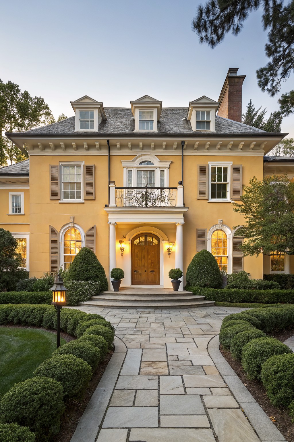 A two-story mustard yellow house with white trim, symmetrical windows, black shutters, a second-floor balcony, columned entry porch, boxwood shrubs, lanterns, and a curved bluestone pathway at dusk.
