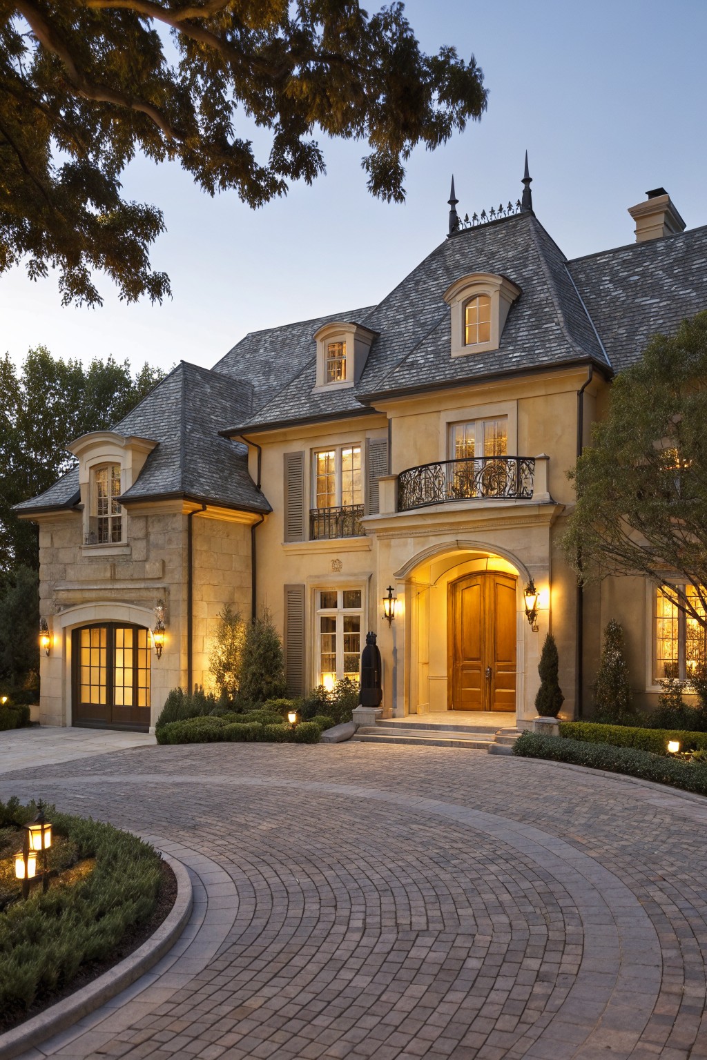 Beige stone house exterior with dark slate roof, arched wooden front door, wrought iron balcony, landscape lights, circular brick driveway, and surrounding trees and shrubs at dusk.